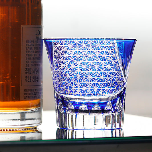 Blue hand cut crystal whiskey glass with geometric facets beside a liquor bottle